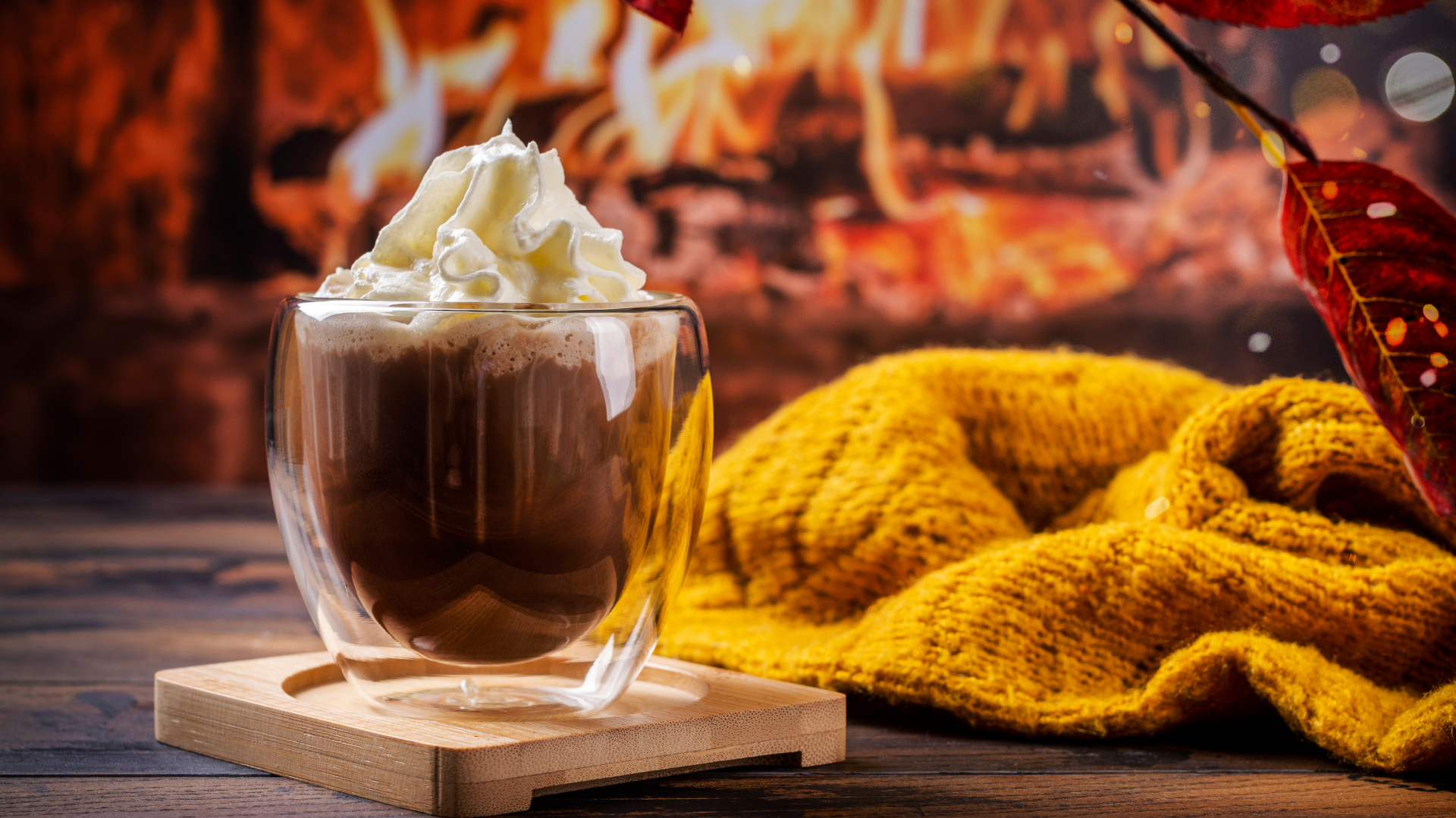 A cozy scene featuring a clear mug filled with a hot drink, topped with whipped cream. The mug sits on a wooden coaster against a blurred background of a warm, crackling fireplace. A soft, yellow knit blanket is draped nearby, and a colorful autumn leaf adds to the ambiance of a chilly fall day indoors.