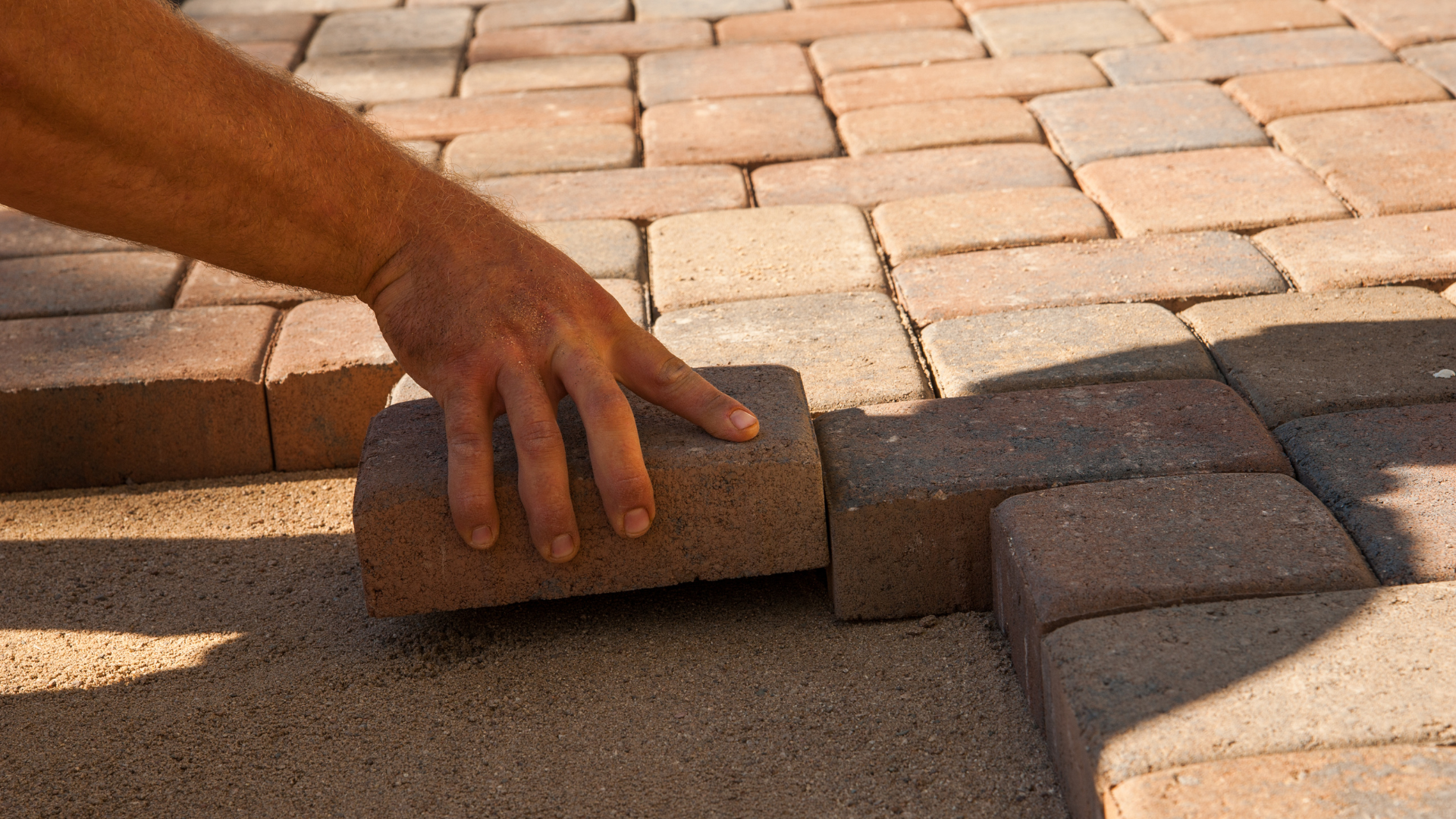 A close-up of a hand placing a brick paver into position on a sandy base, with other pavers already laid out in a pattern beside it.