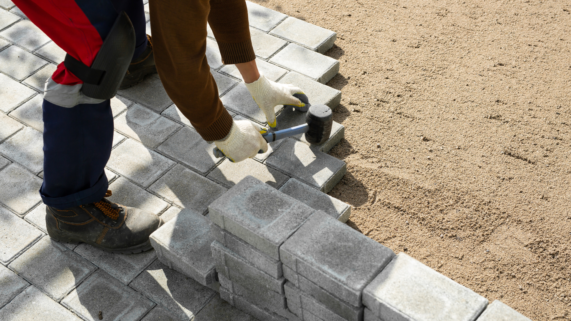 A person laying down stone pavers using a rubber mallet, with a sand base beside the nearly completed pathway.