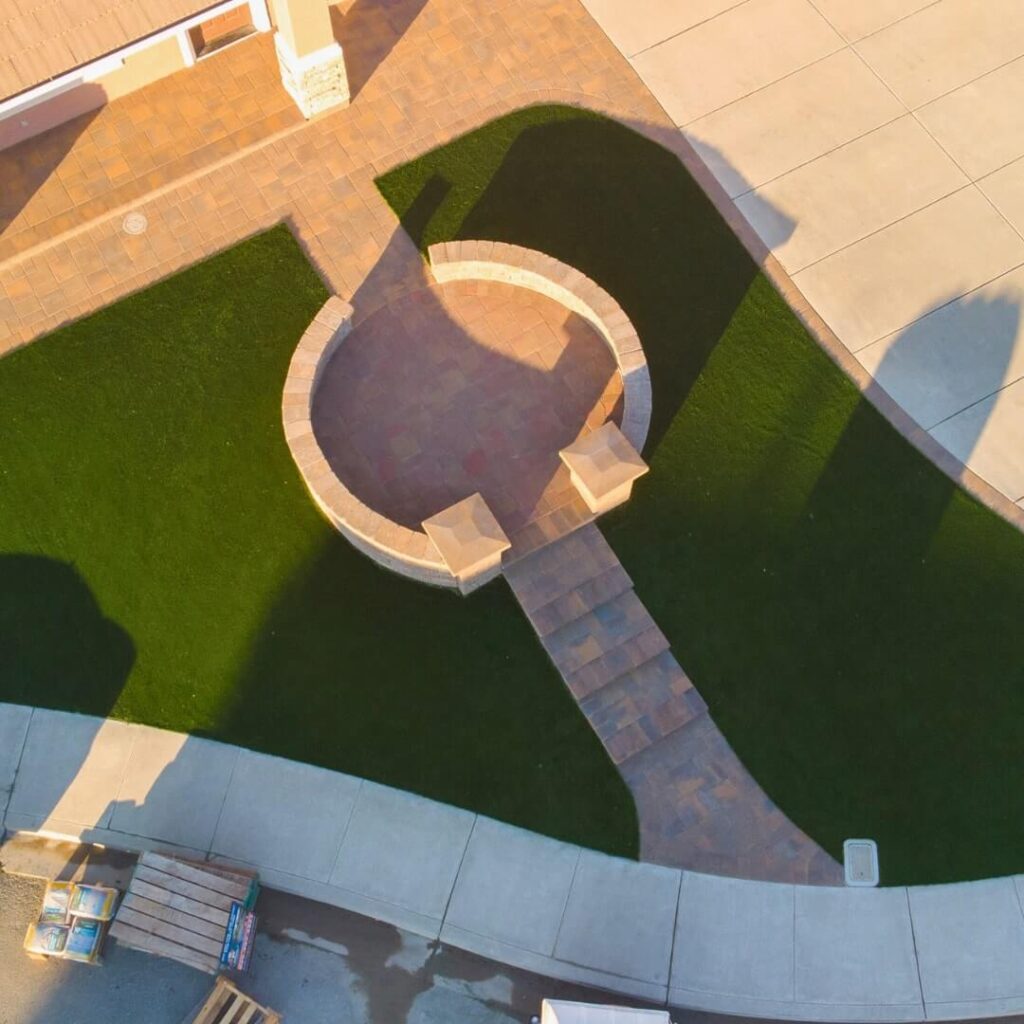 An aerial view showcases a unique garden layout with a bright green lawn. Central to the image is a round, brick-paved feature, possibly a seating or fire pit area, which is connected to the main path by a narrow walkway. The surroundings include various paved sections and seating, with warm sunlight casting elongated shadows.