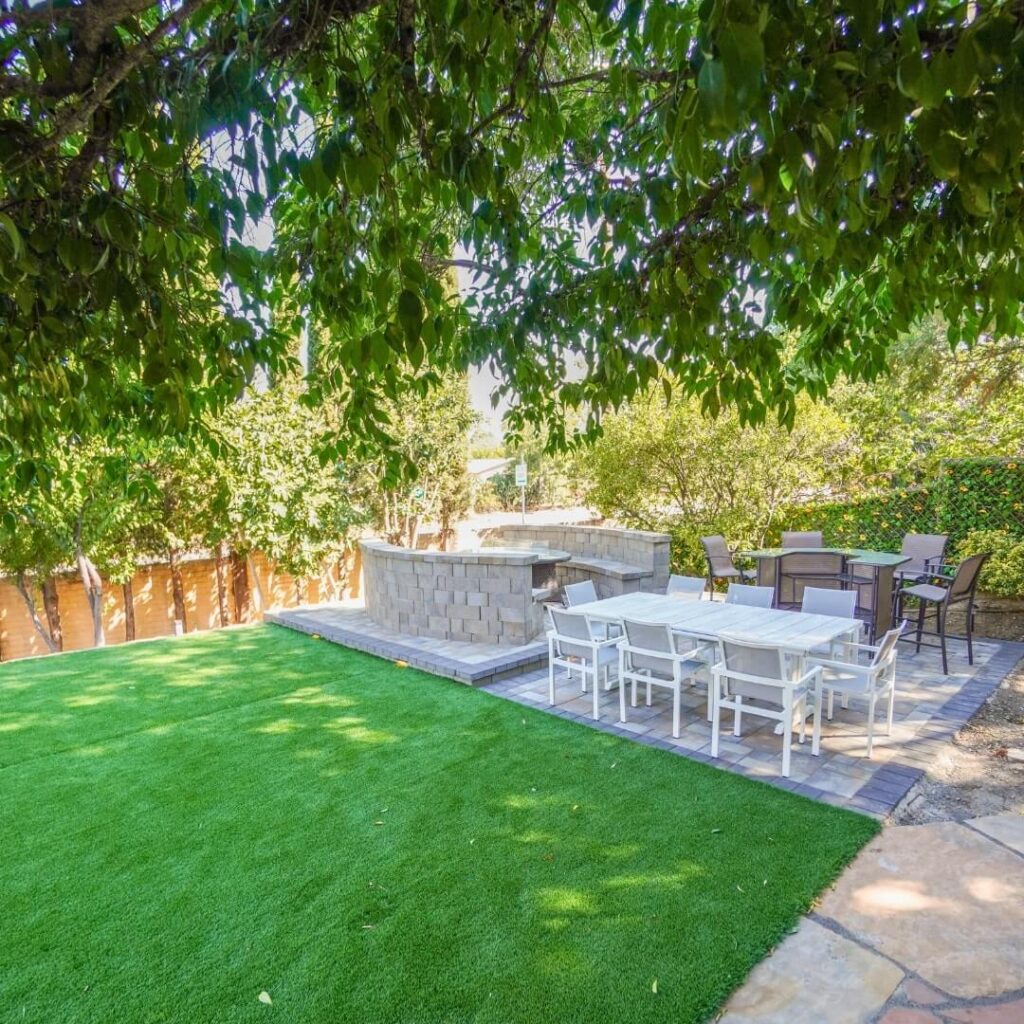A serene backyard setting, highlighted by a vibrant green lawn leading up to an outdoor dining area with a white table and chairs. Adjacent to it is a darker dining set. The scene is framed by a stone barbecue or countertop and lush overhanging branches, adding to the ambiance of a tranquil outdoor space.