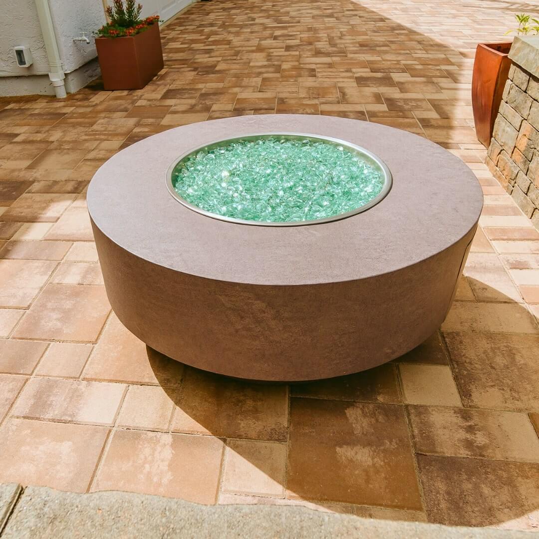 A contemporary round fire pit, shimmering green centerpiece, set on a tiled patio pavers. Adjacent to it are a potted plant and a brick wall structure.