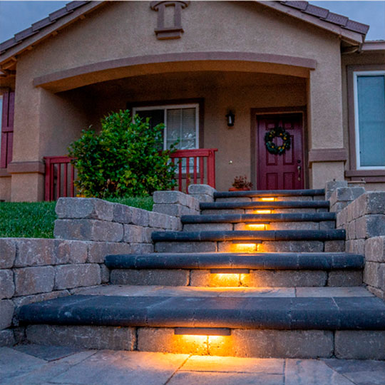 A cozy home entrance featuring stone steps illuminated with warm-toned inset lights leading up to a burgundy door adorned with a wreath. The entrance is sheltered by an arched overhang, complemented by a lush green bush and matching burgundy railings on the side.
