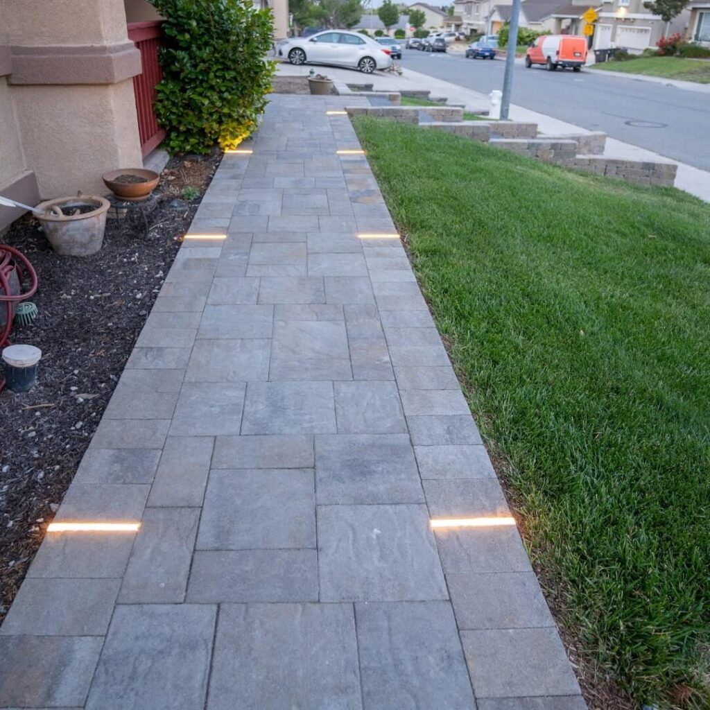 Stone pathway with integrated lighting leading to a residential entrance, with parked cars in the background.