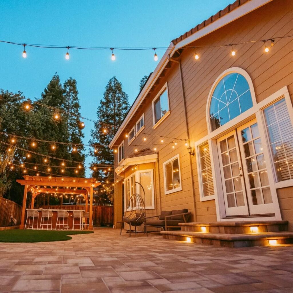 Twilight view of a cozy backyard patio paved with warm string lights, a wooden pergola bar area, and an inviting house facade featuring large arched windows and a comfortable seating area.