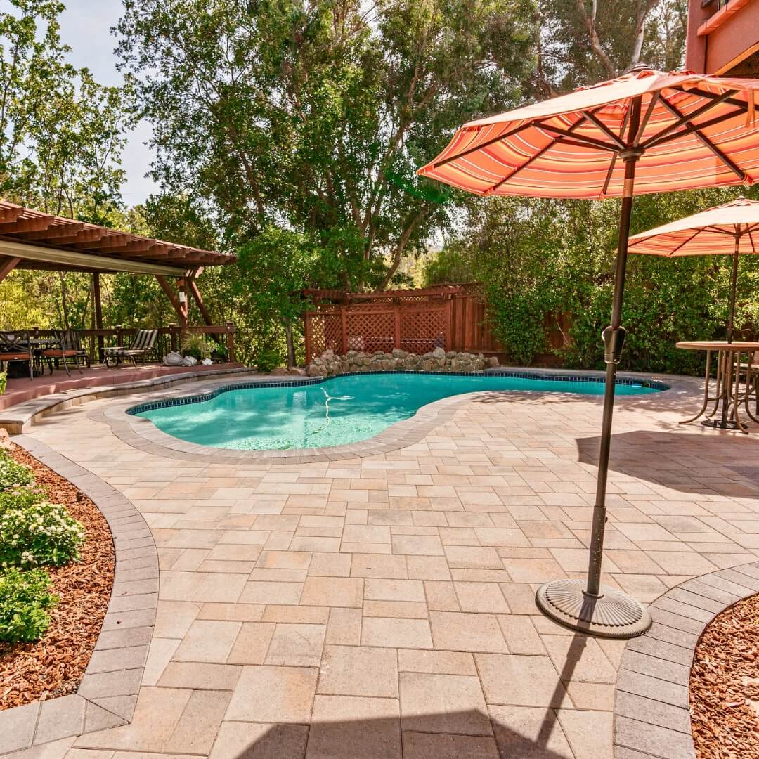 Serene backyard oasis featuring a kidney-shaped pool, stone pavers, shaded seating areas under wooden pergolas, vibrant orange patio umbrellas, and a natural fence of dense trees and shrubs.