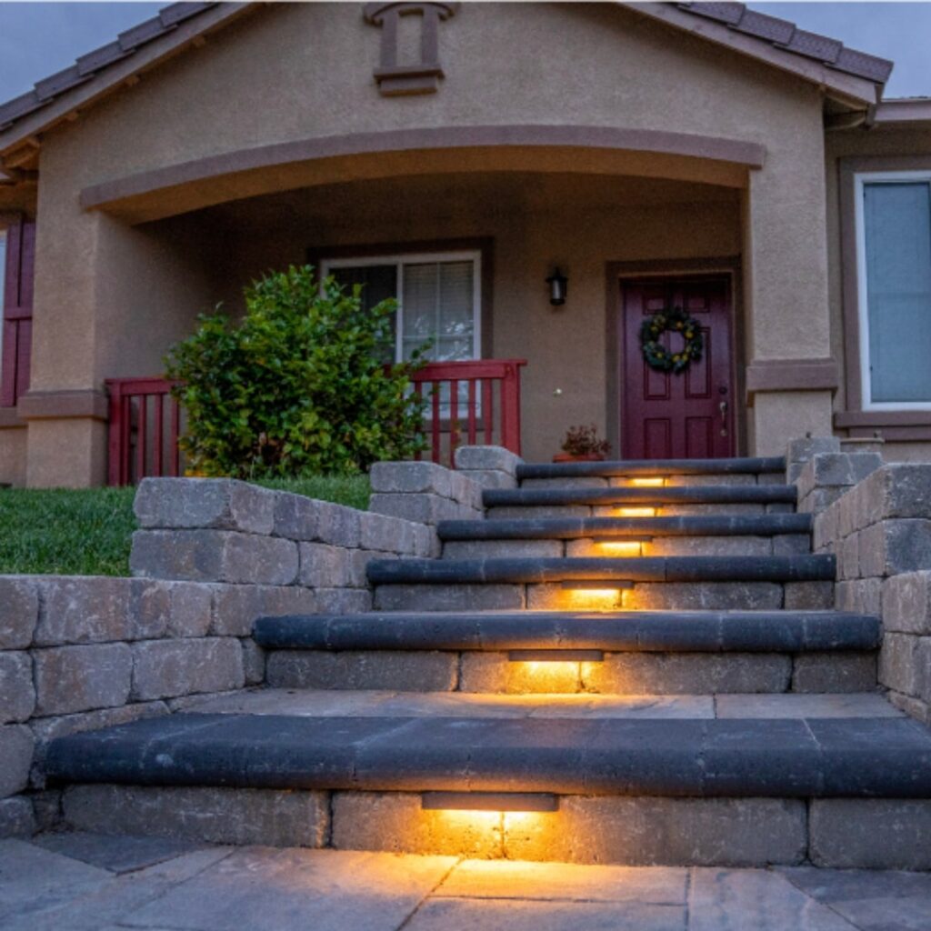 A cozy home entrance featuring stone steps illuminated with warm-toned inset lights leading up to a burgundy door adorned with a wreath. The entrance is sheltered by an arched overhang, complemented by a lush green bush and matching burgundy railings on the side.