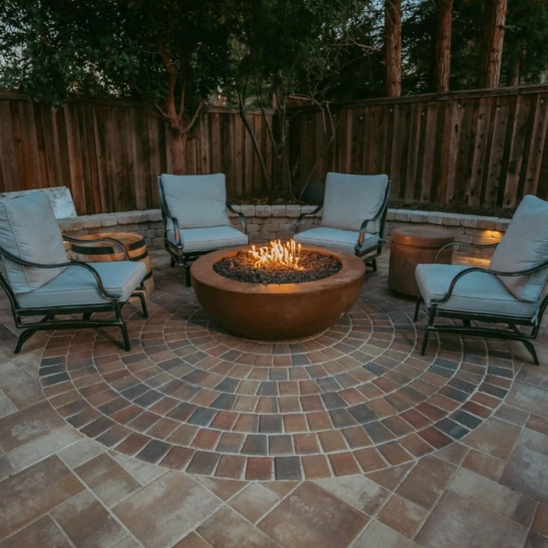 A cozy outdoor setting with a round fire pit in the center, surrounded by four cushioned lounge chairs on a circular brick-patterned patio, enclosed by wooden fences and trees.