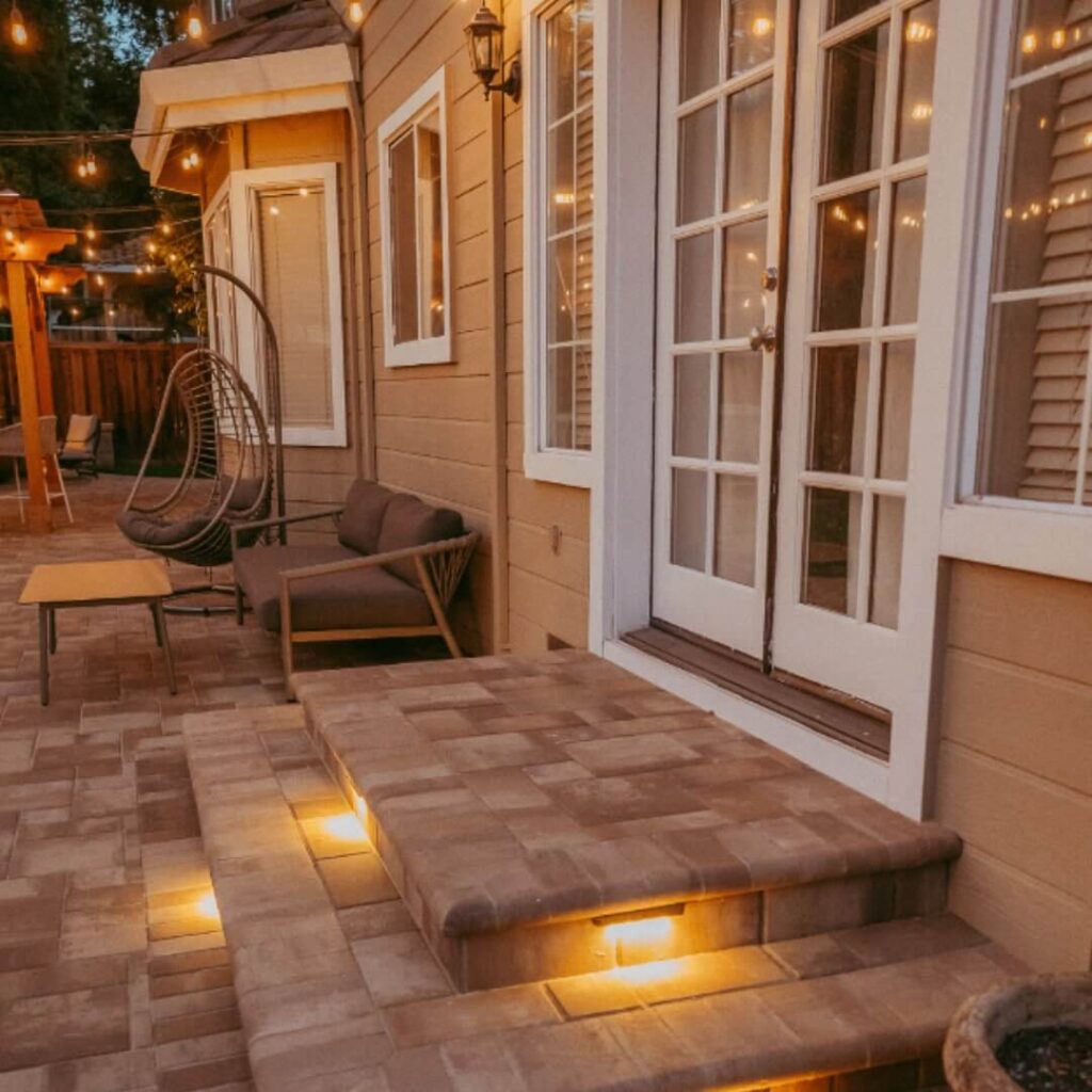 Cozy backyard patio with illuminated steps, hanging egg chair, comfortable seating, and warm string lights near a beige house with large white windows.