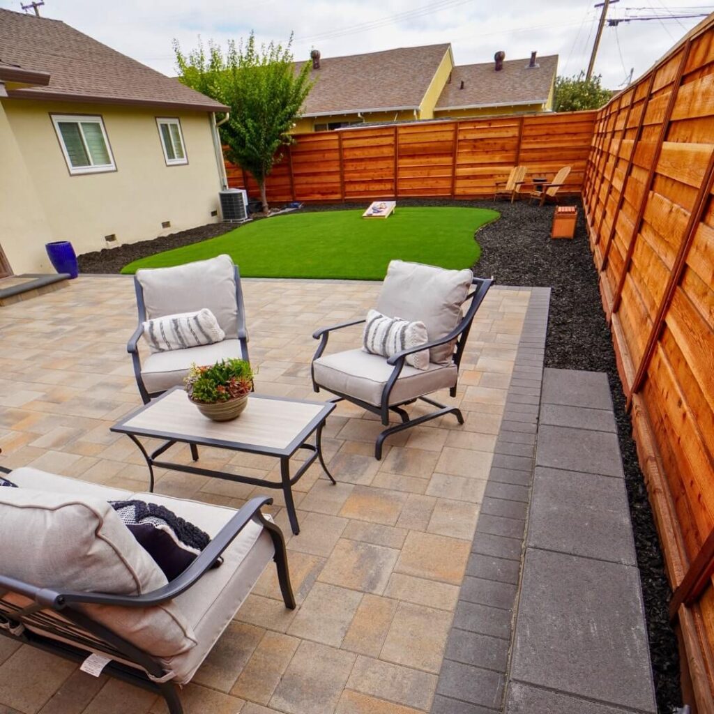Relaxing backyard patio setup with comfortable cushioned chairs and a matching table on multicolored stone pavers, adjacent to a vibrant green artificial lawn and a wooden fence, with a beige home in the background.