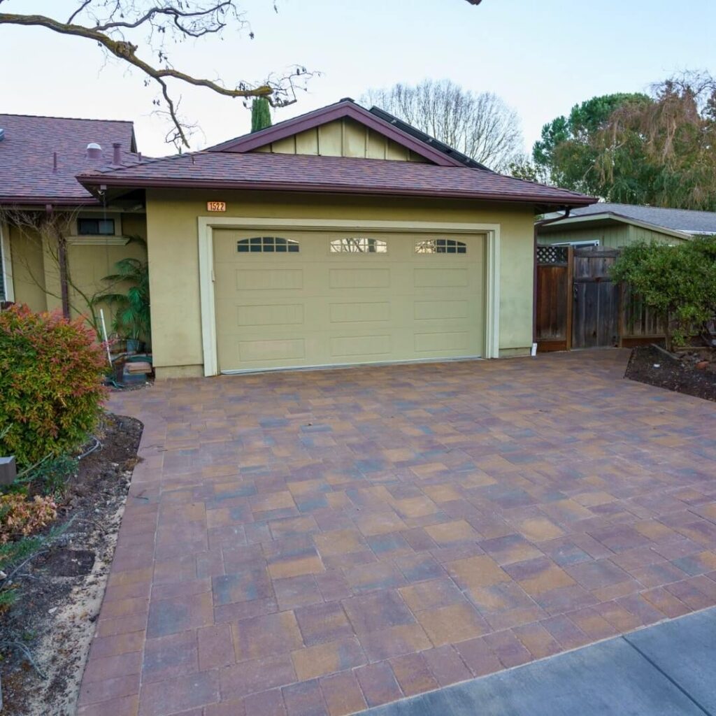 This charming single-story residence boasts a prominent double garage door in a subtle beige tone, complementing the home's soft green walls. Multi-colored pavers form a harmonious driveway, leading the eye to the entrance.