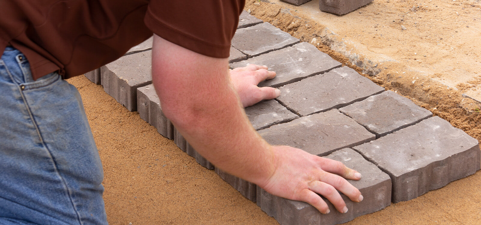 Close-up of a craftsman's hands laying and aligning brick pavers on a sand base, demonstrating attention to detail in patio construction.