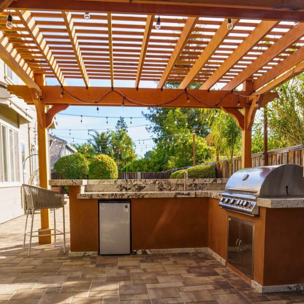 Outdoor kitchen under wooden pergola with stainless steel grill, rustic brick flooring, and string lights, surrounded by greenery.
