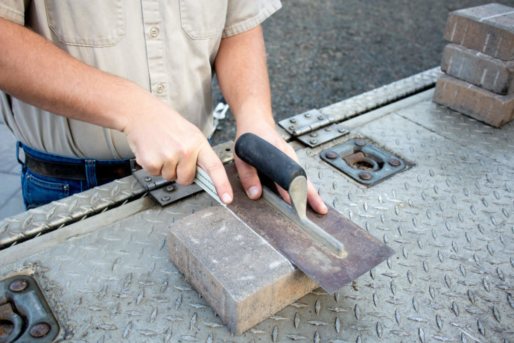 Close-up of a worker's hands using a trowel on a brick, set against a textured metal background, depicting craftsmanship and precision in masonry.