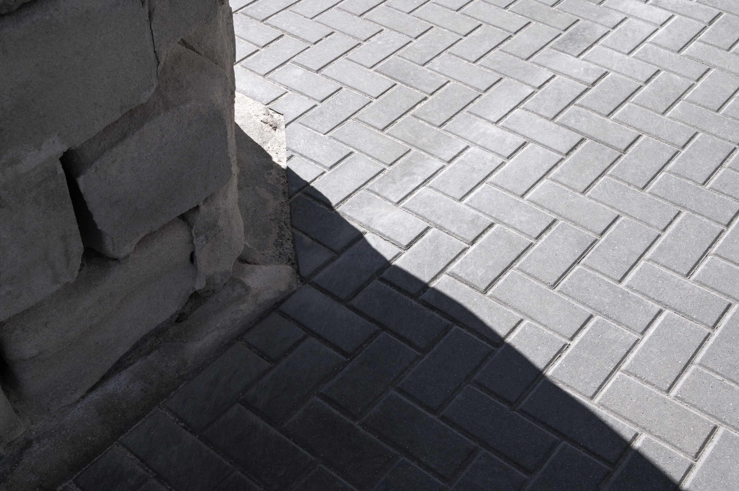 Stack of rough-cut concrete blocks casting a shadow, adjacent to a neatly laid square-patterned pavement, emphasizing contrast in construction materials.