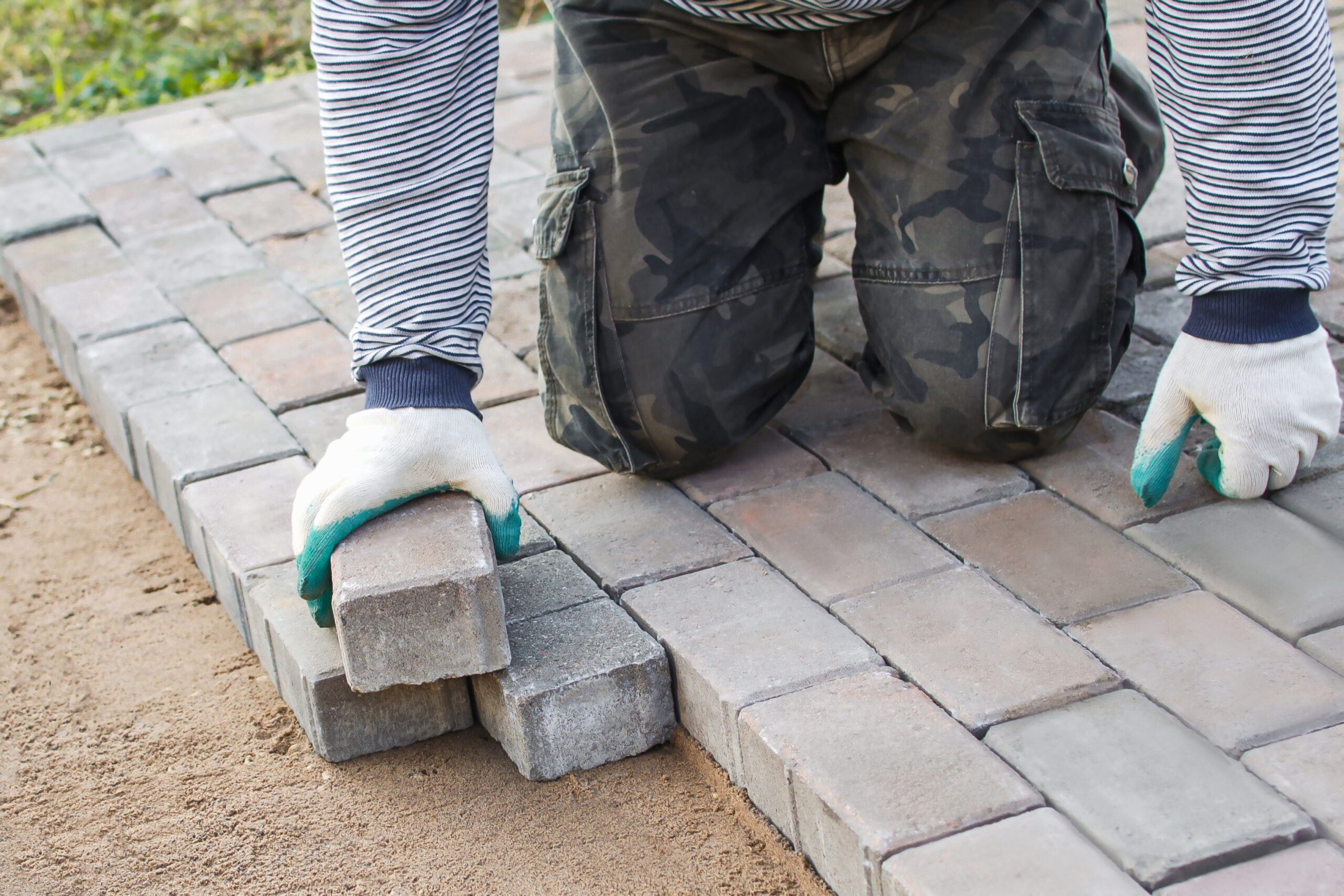 Worker in striped shirt and camo pants placing paving stones on a pathway outdoors.