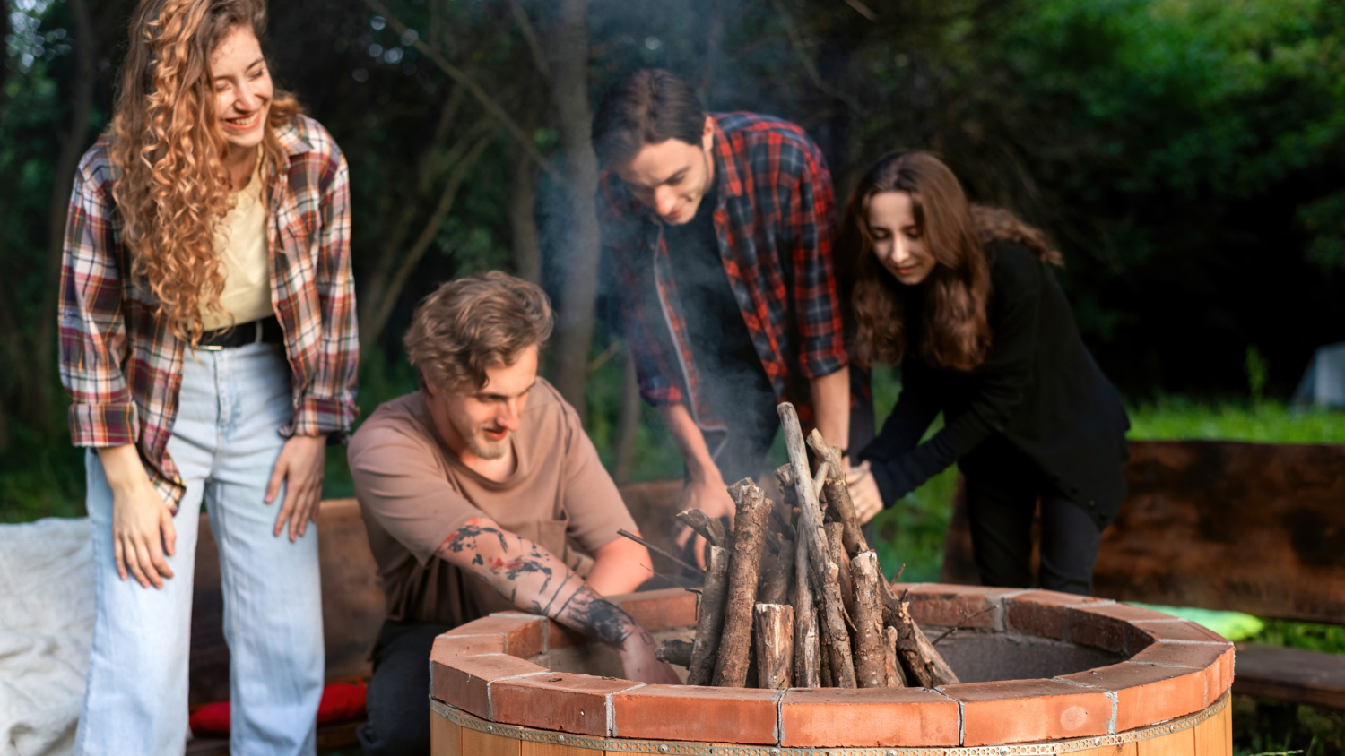 A group of four friends arranging wood in a brick fire pit in a green backyard, with one man kneeling and others standing, smoke rising, in casual attire, enjoying outdoor activities together.