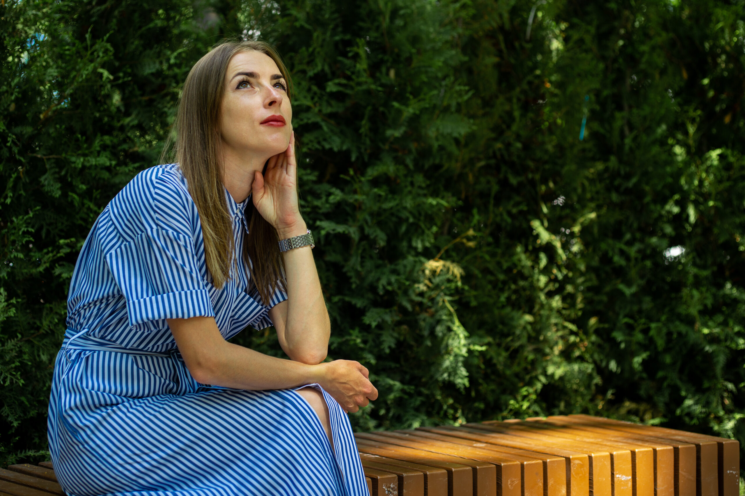 A woman in a blue and white striped dress sitting on a park bench, looking thoughtfully upwards with her hand on her cheek, surrounded by lush greenery