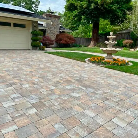 A beautifully paved driveway complements the house's facade with stone accents. To the side, a classic multi-tiered fountain is surrounded by vibrant yellow flowers, all of which is enclosed by a manicured lawn and tall trees in the background. The property exudes a sense of serenity and elegance.