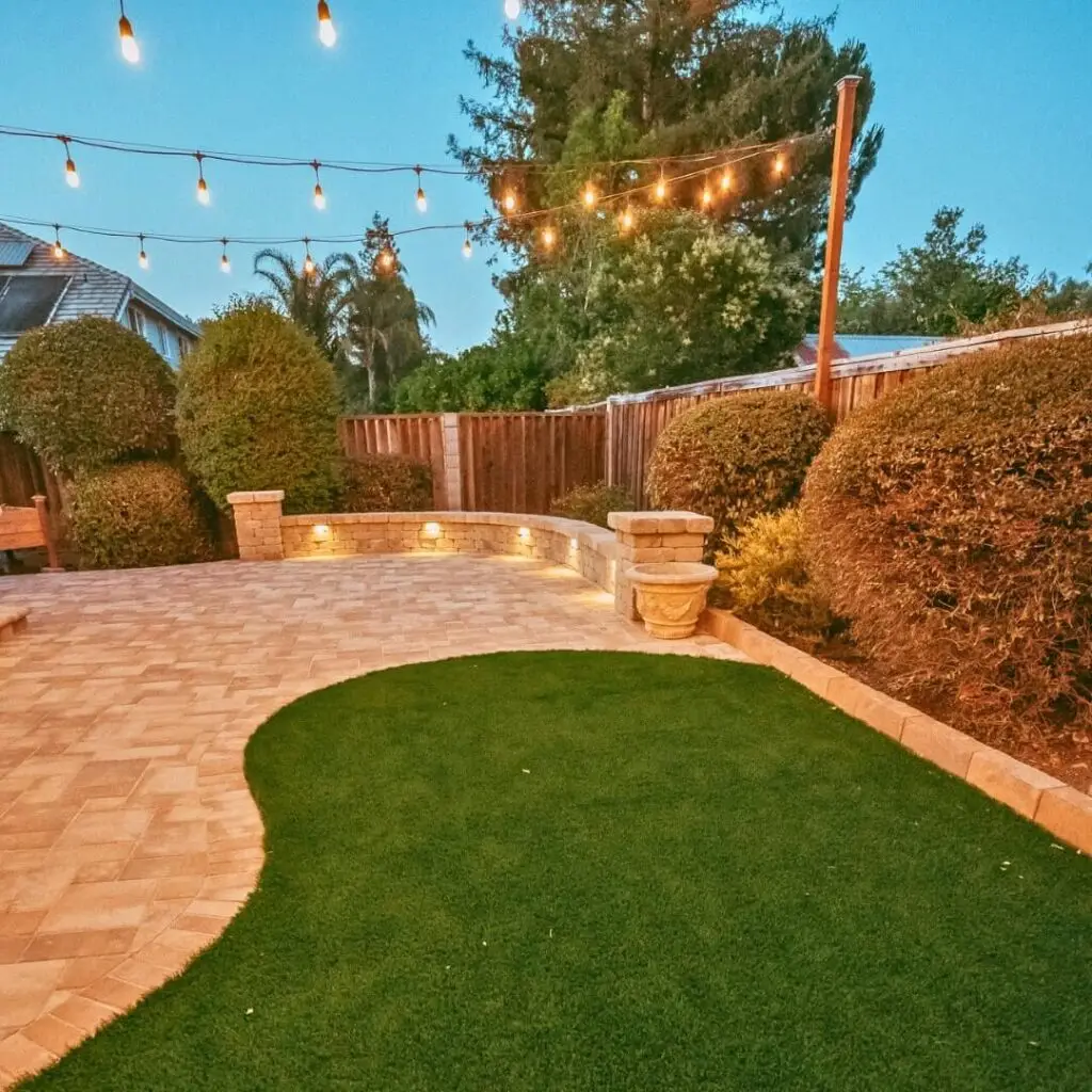 Cozy backyard design with illuminated string lights overhead, elegant stone pavers, lush green artificial turf, and decorative stone planters, all bordered by mature shrubs and a wooden fence during dusk.