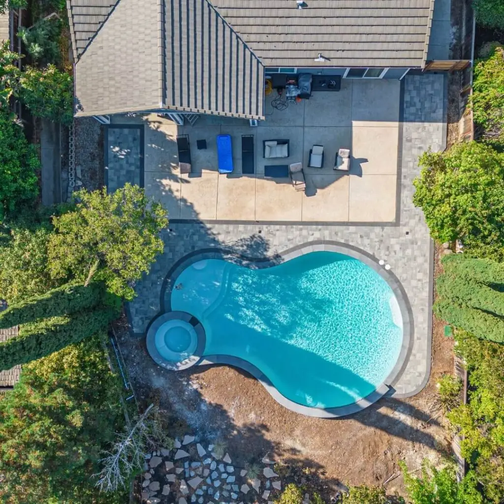 Aerial view of a modern home with a unique kidney-shaped swimming pool, surrounded by lush greenery, a paved patio with outdoor furniture, and a shaded pergola area, captured on a bright sunny day.