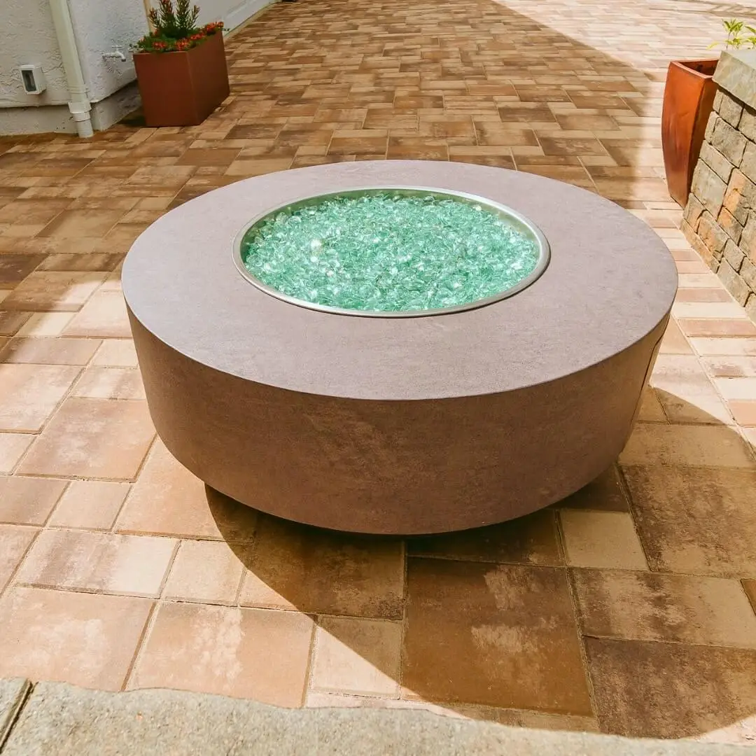 A contemporary round fire pit, shimmering green centerpiece, set on a tiled patio pavers. Adjacent to it are a potted plant and a brick wall structure.
