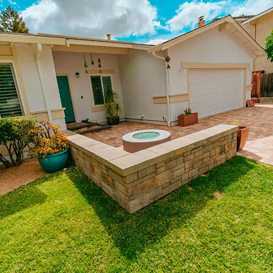 A well-laid out patio area featuring a blend of rich-toned pavers that create a harmonious contrast with the lush green lawn. The pavers extend to form an elevated platform, which houses a decorative circular element, all bordered by a sturdy brick wall that adds dimension and texture to the space.