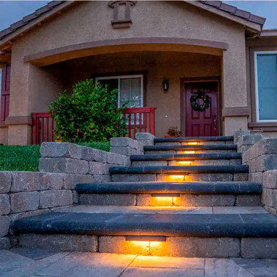 A cozy home entrance featuring stone steps illuminated with warm-toned inset lights leading up to a burgundy door adorned with a wreath. The entrance is sheltered by an arched overhang, complemented by a lush green bush and matching burgundy railings on the side.
