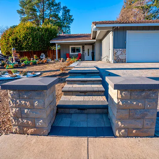 Inviting home exterior featuring a well-manicured garden, modern stone paver pathway with symmetrical steps, flanked by robust stone pillars all made of pavers, leading to a cozy house with a gray roof, white sidings, and an attached garage.