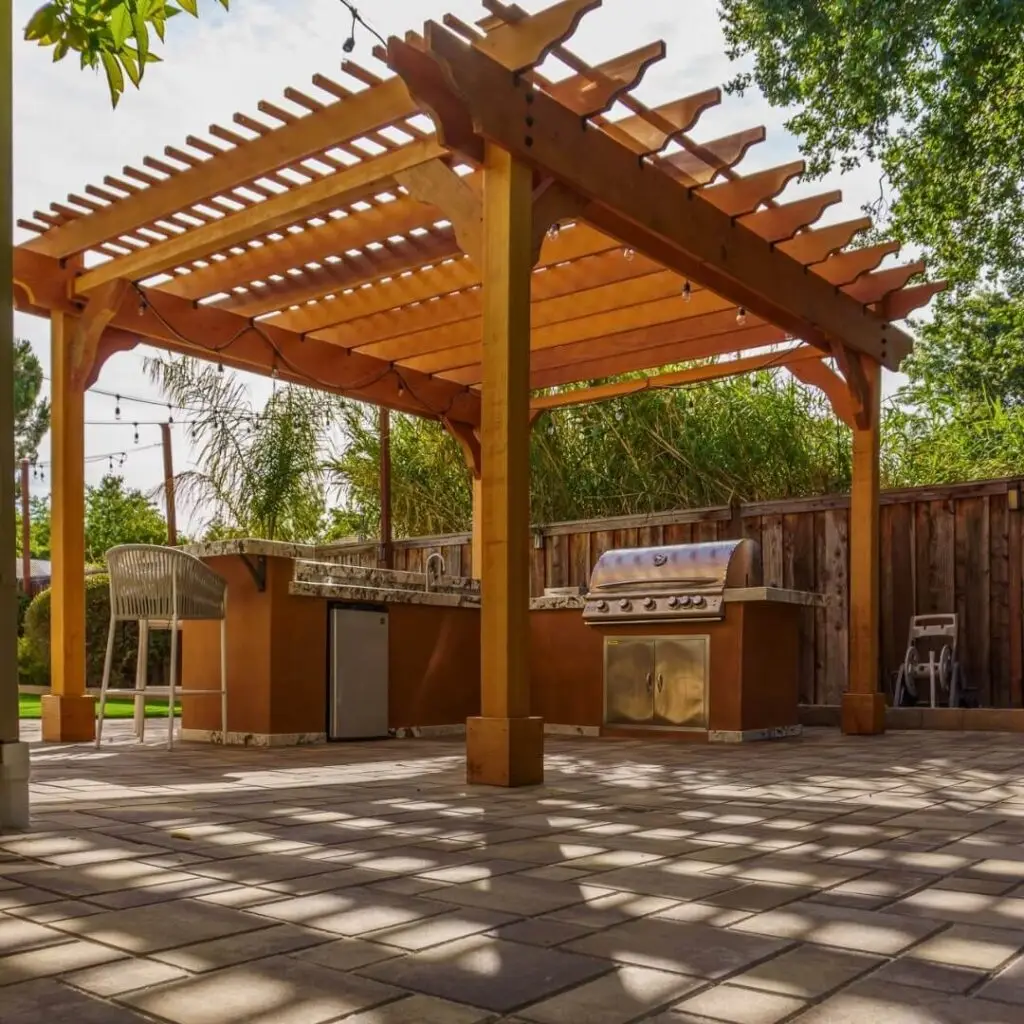 Outdoor kitchen under wooden pergola with stainless steel grill, rustic brick flooring, and string lights, surrounded by greenery.