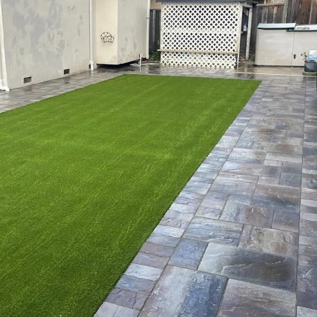 Neatly manicured green artificial lawn adjacent to a patterned gray stone patio, with a glimpse of a light gray house exterior and white lattice screen in the background.