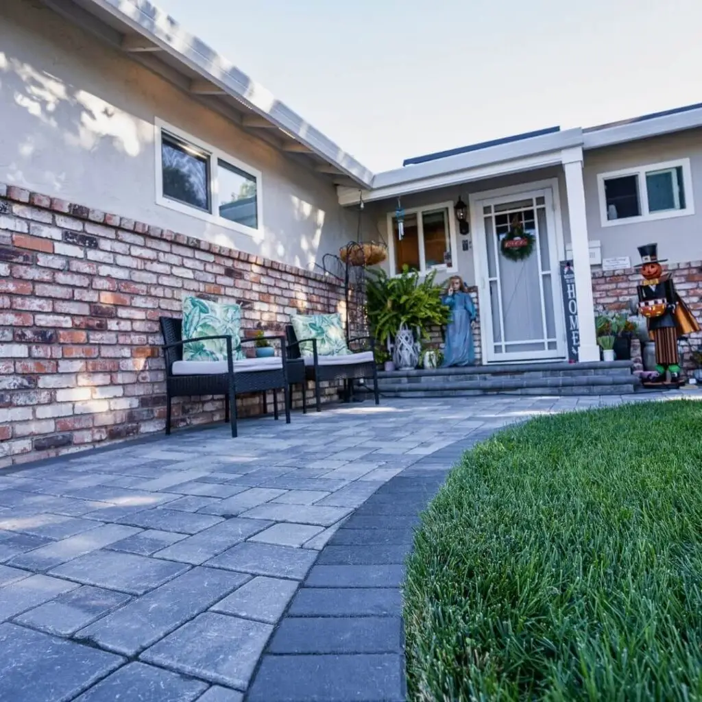 Charming outdoor patio with brick wall detailing, complemented by dark stone pavers, lush green lawn, and inviting entrance adorned with festive decorations and tropical-themed seating area.