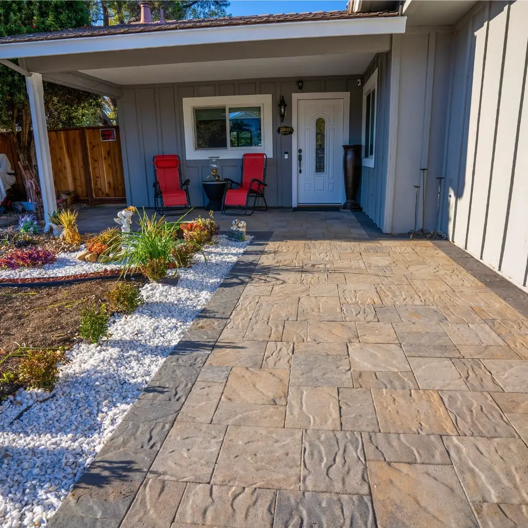 Stone-patterned paver patio leading to a cozy home entrance, accentuated by vibrant red chairs, white ornamental gravel, and a diverse mix of decorative plants under a sunlit sky.