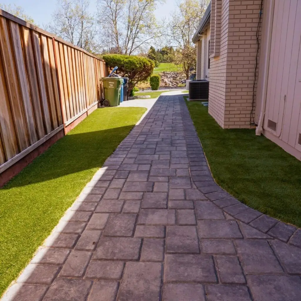 Well-maintained side yard featuring a path laid with rectangular stone pavers, bordered by a vibrant green artificial lawn on one side and a wooden fence on the other, leading towards a serene garden area with a beige brick home exterior visible to the right.