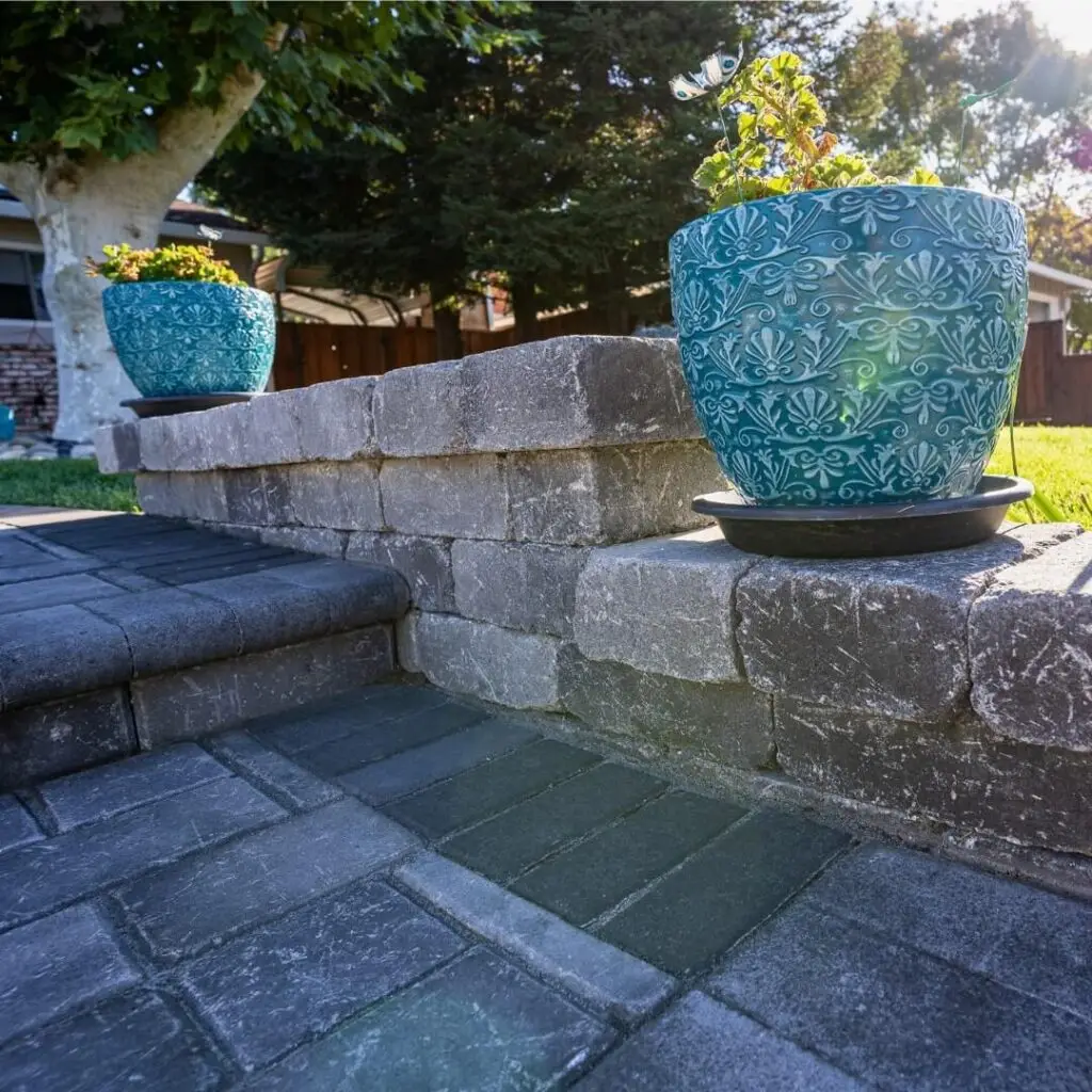 Decorative blue ceramic planter with intricate floral design, placed on a grey stone wall, surrounded by paved patio, ideal for garden decor and outdoor aesthetics.