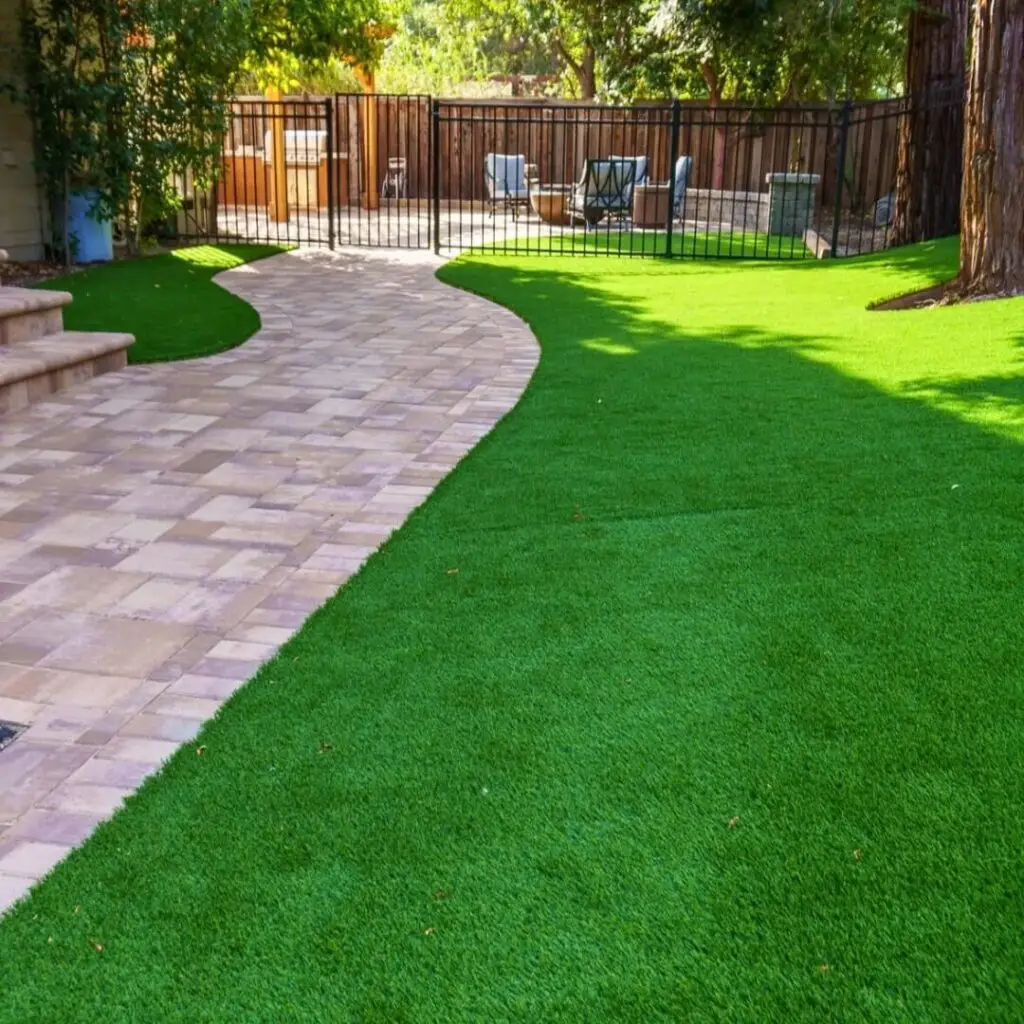 Lush green artificial lawn adjacent to a curving stone-paved pathway leading to a fenced outdoor seating area with comfortable furniture, surrounded by mature trees providing shade.