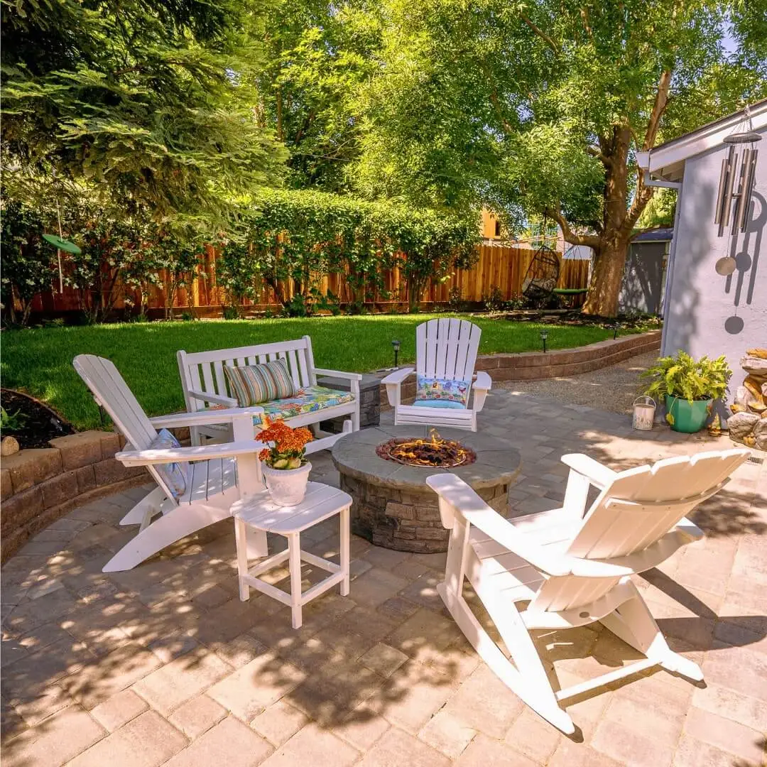 Sunlit backyard patio featuring white Adirondack chairs, decorative cushions, a stone fire pit, potted greenery, interlocking pavers, and a wooden fence backdrop.