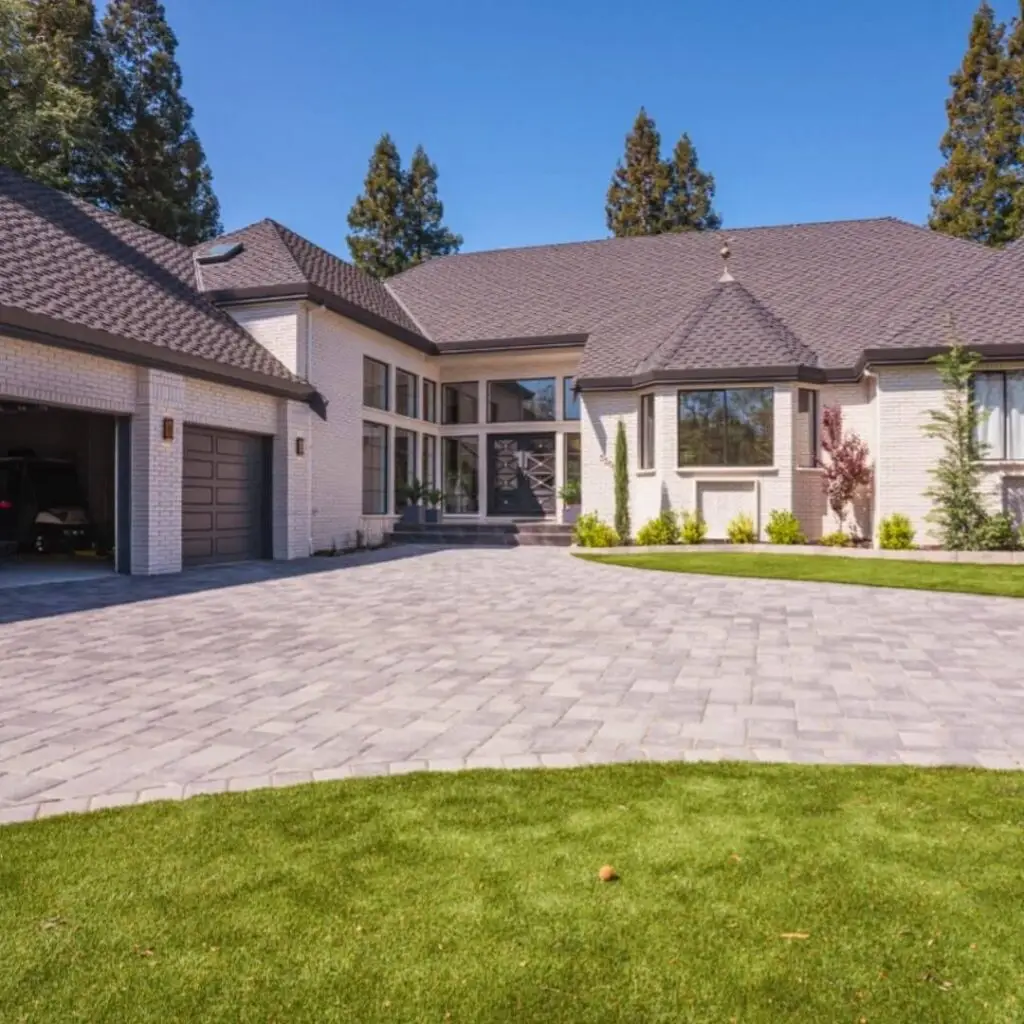 This expansive property showcases a luxurious home with a blend of modern and classic architectural elements. The dark roofs contrast beautifully with the white brick walls, while large windows and glass doors offer ample natural light and views of the surroundings. The wide driveway, paved with light-colored bricks, extends to a spacious garage. Lush green grass and strategically placed plants add a touch of nature and freshness to the meticulously designed residence, creating an inviting and grand atmosphere.