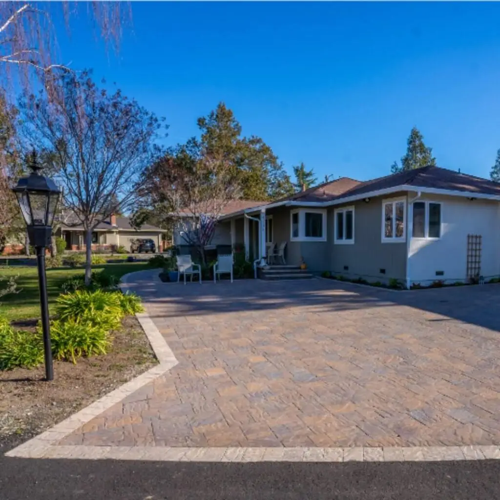 Spacious suburban home with taupe exteriors, accompanied by a beautifully paved driveway. The front yard features manicured greenery, a vintage lamp post, and patio furniture, all under a clear blue sky.