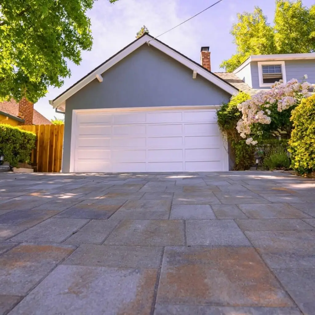 Sunny view of a detached garage painted in dark grey, with a white door, set against a brick home. A patterned stone driveway leads up to it, bordered by lush greenery and blooming white flowers.