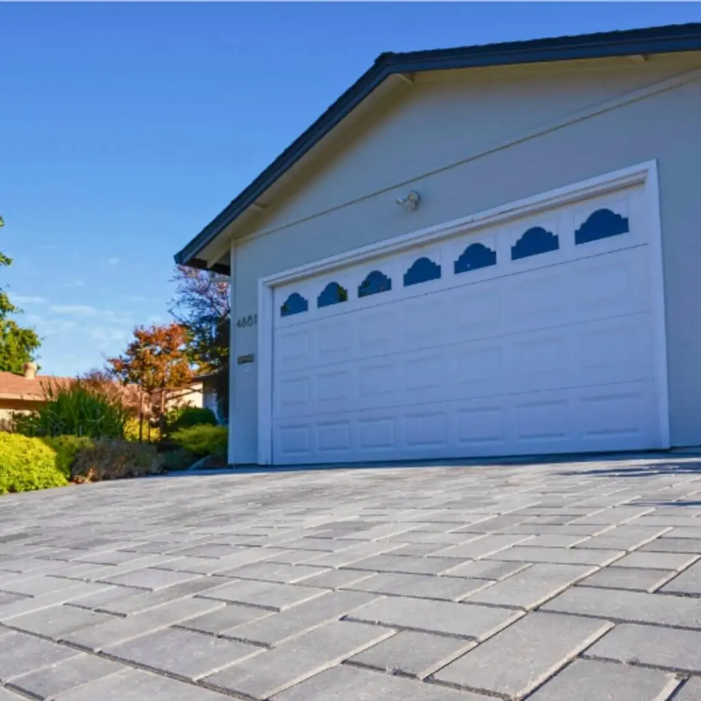 Exterior view of a modern home with white garage door featuring arched window designs, set against a clear blue sky. Grey brick-paved driveway leads to address 4601, complemented by landscaped greenery.